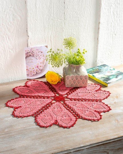 Rundes gehäkeltes Deckchen in Rosé mit rotem Rand auf hellem Holztisch, daneben Vase und Blume