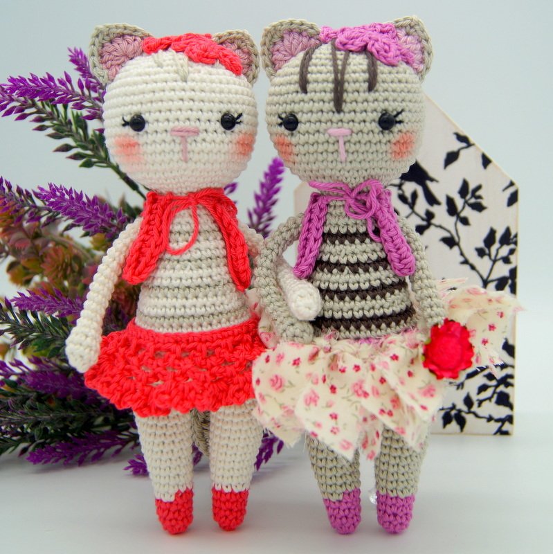 Zwei gehäkelte Amigurumi-Katzenpuppen nebeneinander, eine mit rotem Rock und Halstuch, die andere gestreift mit geblümtem Rock und violettem Halstuch
