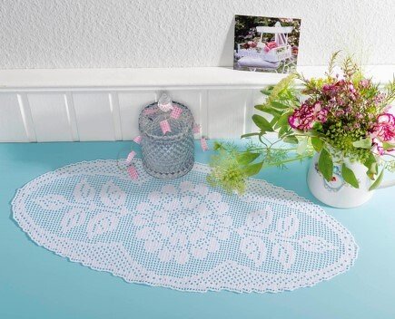 Ovales gehäkeltes Deckchen mit Blattmuster auf türkisfarbenem Untergrund, daneben eine Glasdose und ein Blumenstrauß