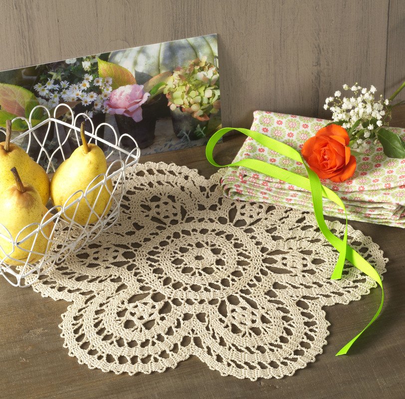Beiges gehäkeltes Deckchen mit filigranem Blumenmuster auf Holztisch, neben Birnen und Blumen.