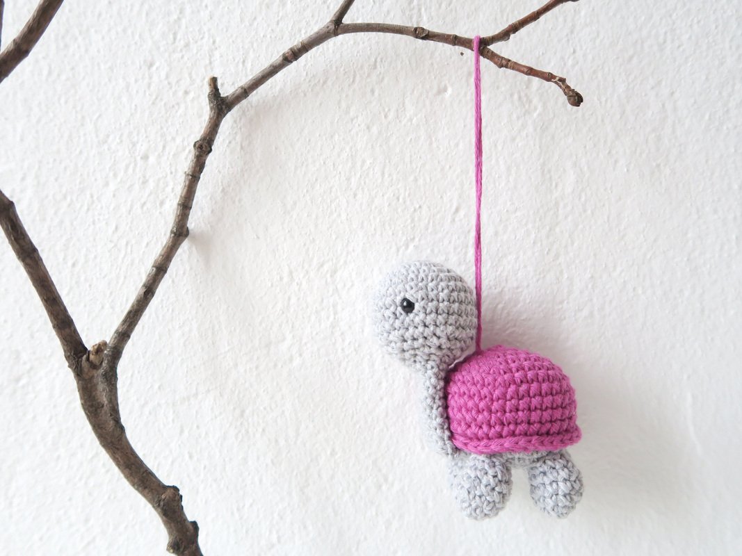 Gehäkelte Amigurumi-Schildkröte in Grau mit pinkem Panzer, an einem Ast hängend vor weißer Wand.