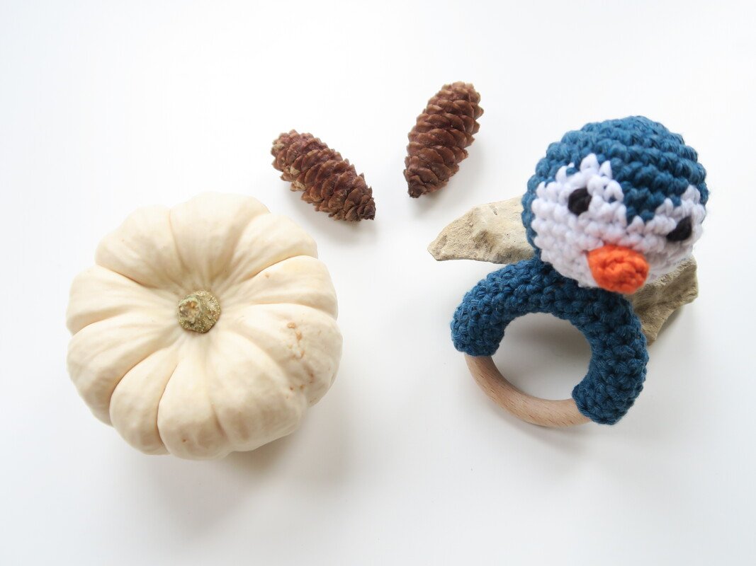 Gehäkelte blaue und weiße Pinguin-Rassel mit Holzring neben hellem Zierkürbis und zwei Tannenzapfen auf weißem Untergrund.