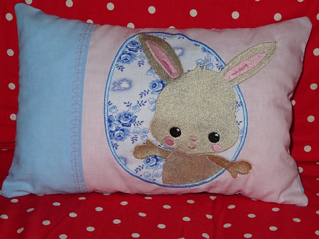 Rosa-hellblaues Kissen mit Hasen-Applikation im ovalen, blau geblümten Rahmen auf rotem Stoff mit weißen Punkten.