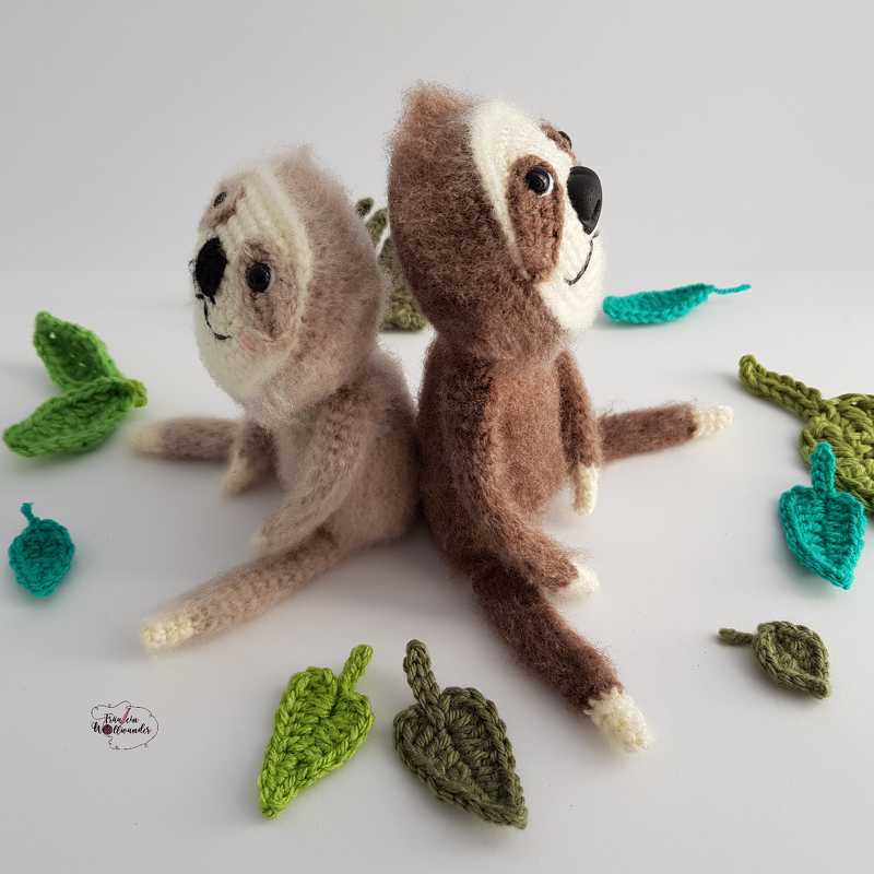 Zwei gehäkelte Amigurumi-Faultiere, hell und dunkel, sitzen Rücken an Rücken auf weißem Hintergrund mit gehäkelten grünen Blättern.