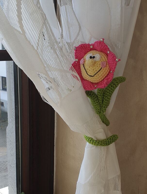 Gehäkelter Blumen-Raffhalter in Pink mit gelbem Smiley-Gesicht und grünen Blättern an weißem Vorhang