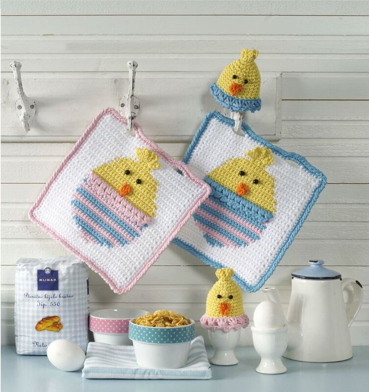 Gehäkelte Topflappen mit Kükenmotiv und passende Eierwärmer in Pastellfarben an Haken und auf einer Küchentheke.