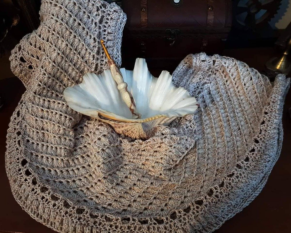 Nahaufnahme eines beigen, gehäkelten Loop-Schals mit Muschel und Häkelnadel auf dunklem Untergrund.