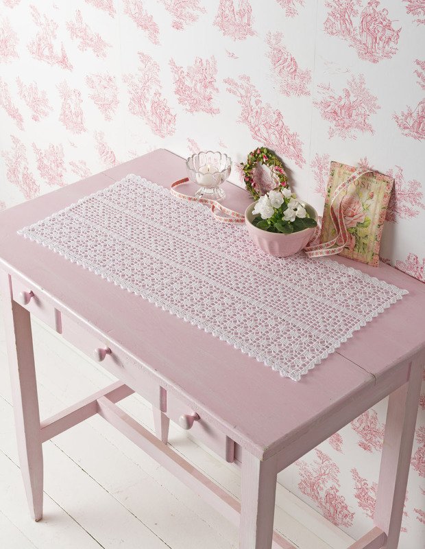 Rechteckiger weißer Häkel-Tischläufer mit Muschelrand auf rosa Holztisch, dekoriert mit Schale und Blumen