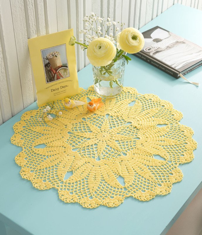 Gelbes rundes gehäkeltes Deckchen mit Blumenmuster auf türkisfarbenem Tisch, Vase mit Blumen und kleinen Dekofiguren