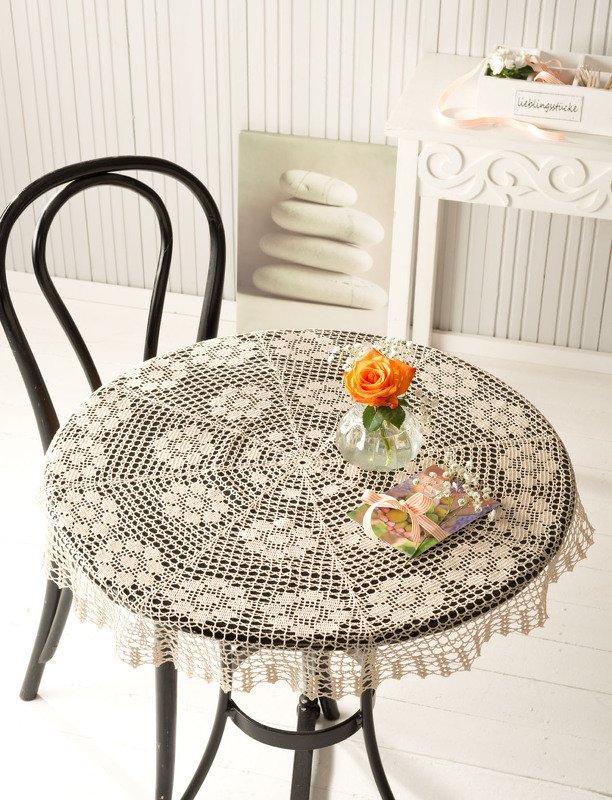 Rundes gehäkeltes Spitzen-Deckchen auf kleinem Tisch mit Glasvase und orangefarbener Rose