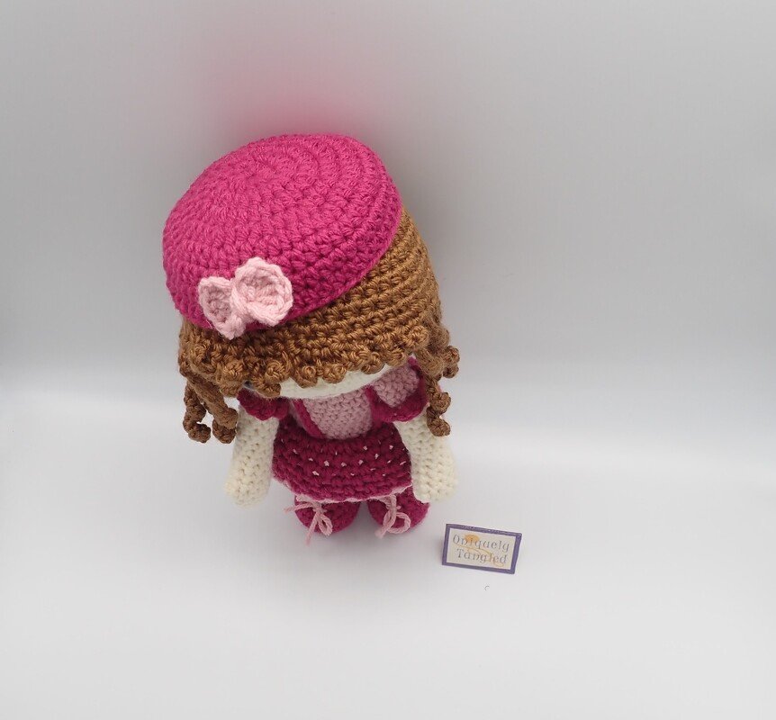 Crocheted amigurumi doll with pink beret, curly brown hair, and textured skirt viewed from an elevated angle