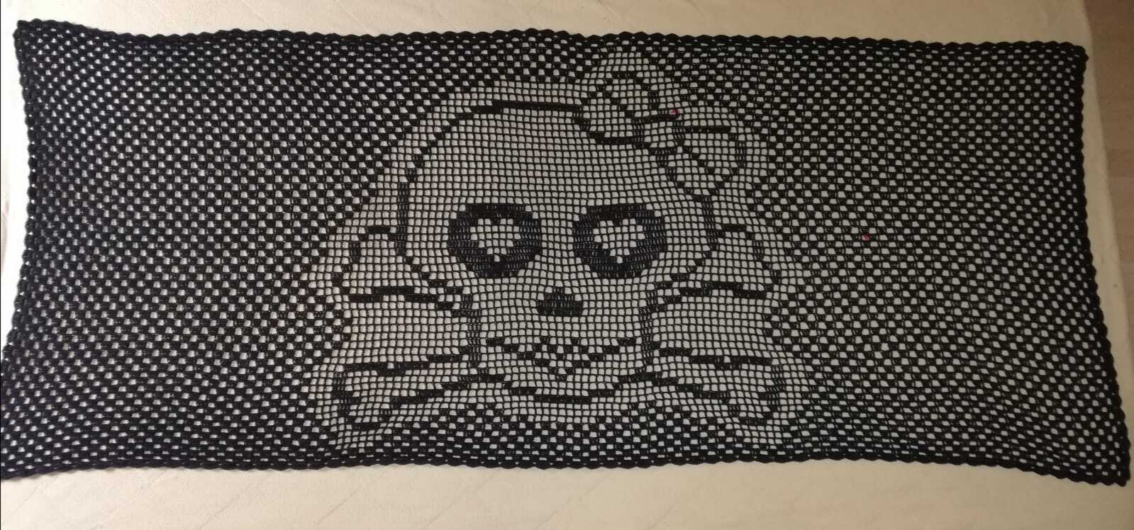 Rechteckiges schwarzes Filet-Häkeltuch mit Totenkopf- und gekreuzten Knochen-Motiv auf hellem Untergrund.