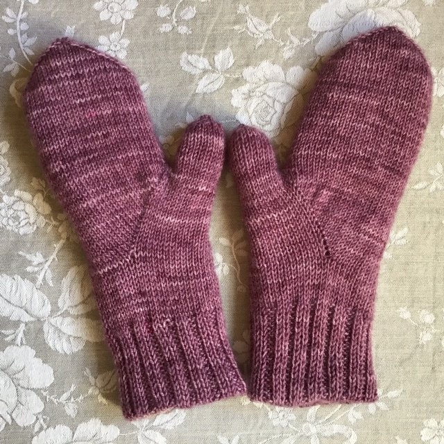 Zwei handgestrickte Fäustlinge mit Daumen in meliertem Rosa-Lila auf floralem Stoff liegend.