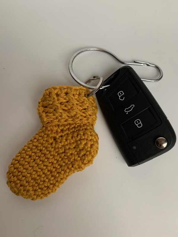 Gehäkelter gelber Babyschuh‑Schlüsselanhänger neben Autoschlüssel auf weißem Hintergrund