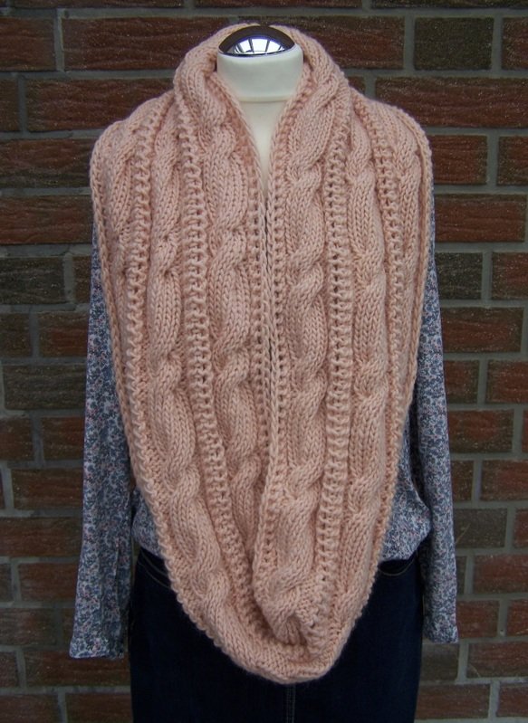 Zartrosa gestrickter Loopschal mit Zopfmuster auf Schneiderpuppe vor Backsteinwand.