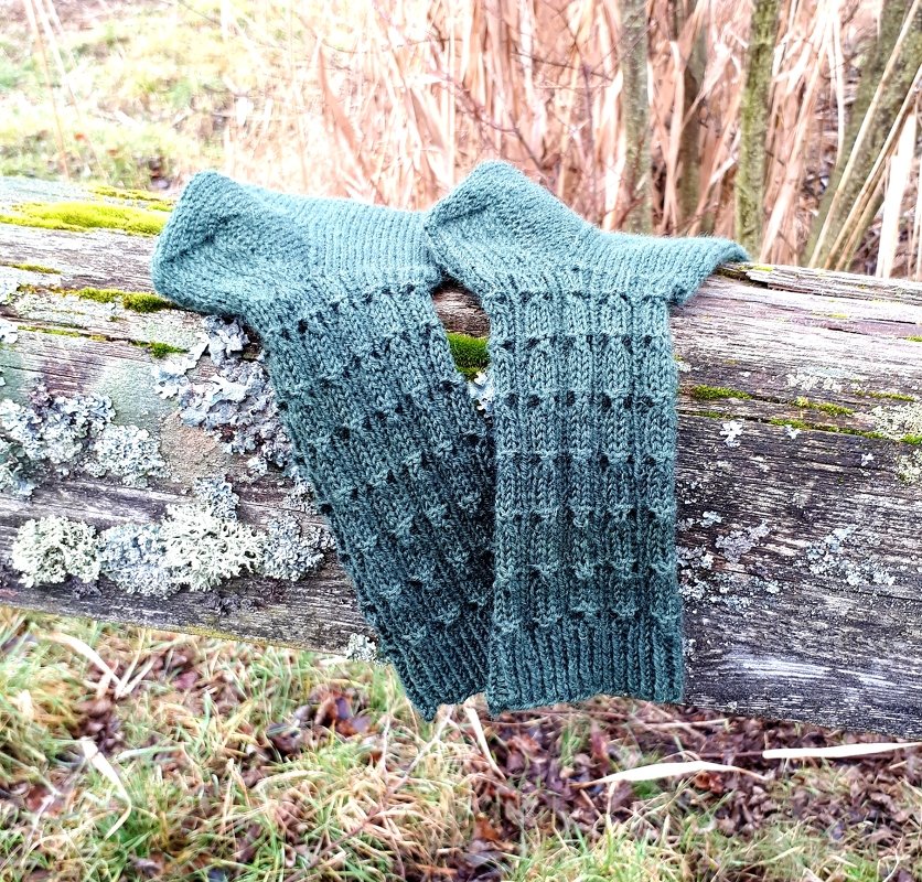 Zwei dunkelgrüne gestrickte Beinstulpen mit strukturiertem Muster auf einem moosbewachsenen Holzstamm.