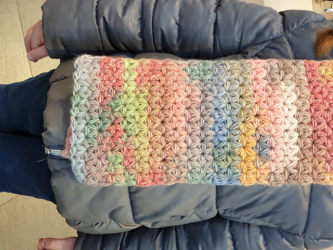 Bunter gehäkelter Kinderschal in Pastellfarben mit Sternchenmuster, getragen zu einer grauen Winterjacke.