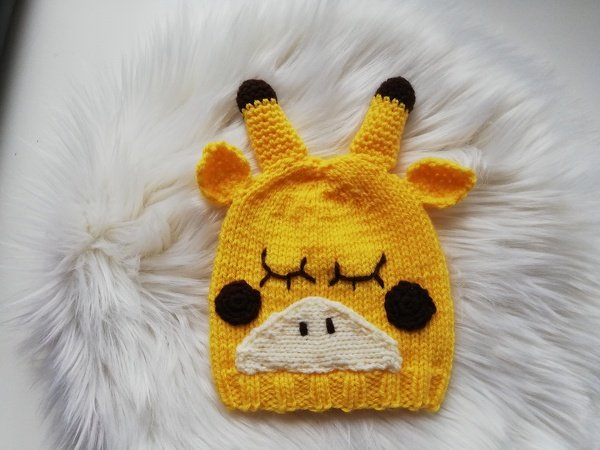 Gestrickte gelbe Babymütze in Giraffenform mit Ohren, Hörnern, geschlossenen Augen und Schnauze auf weißem Kunstfell.