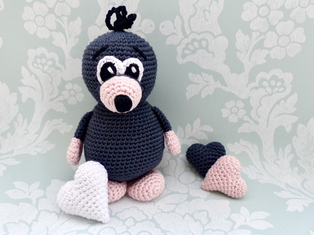Gehäkelte dunkelgraue Amigurumi-Maulwurf-Figur mit rosa Schnauze, rosa Pfoten und drei gehäkelten Herzen in Weiß, Rosa und Dunkelgrau