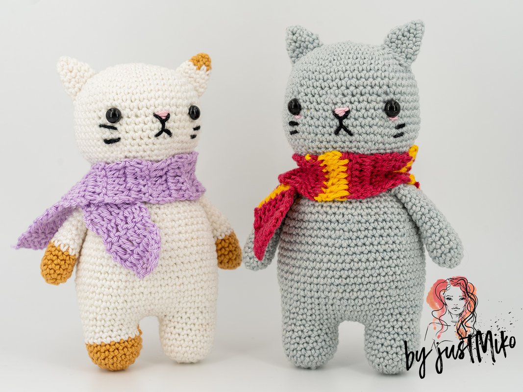 Zwei gehäkelte Amigurumi-Katzen, eine weiße mit lila Schal und braune Akzente, eine graue mit rot-gelbem Schal.