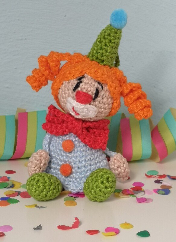 Gehäkelter Clown-Amigurumi mit orangen Zöpfen, grüner Mütze und roter Fliege sitzt zwischen Konfetti