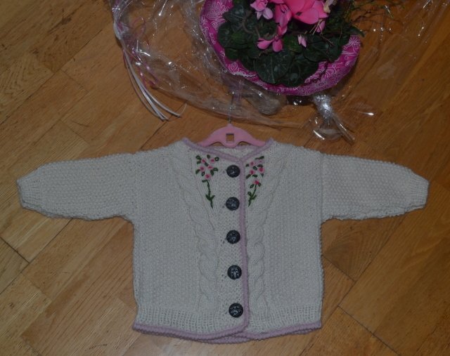 hellbeige gestrickte Babyjacke mit Blumenstickerei und dunklen Knöpfen auf Holzboden