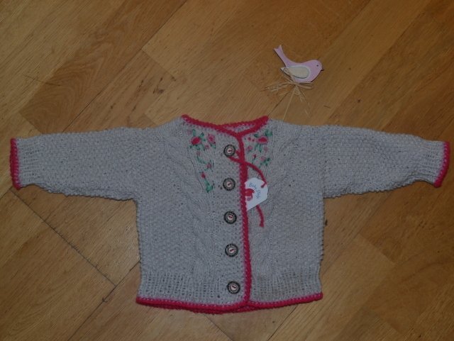 Hellgraue gestrickte Babyjacke mit rosa Zierkante und Blumenstickerei auf Holzboden