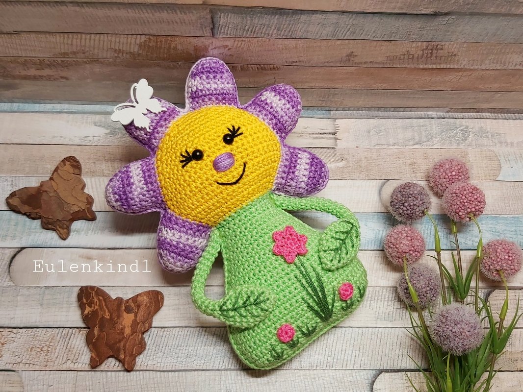 Gehäkelte Amigurumi-Blume mit lächelndem Gesicht, lila Blütenblättern und grünem Körper mit applizierten Blüten