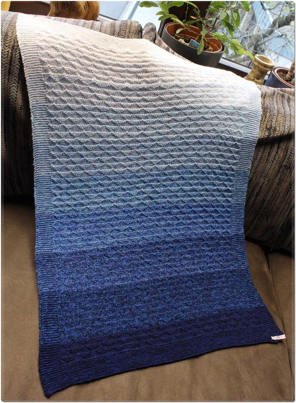 Gestrickte Babydecke mit Farbverlauf von Weiß zu Dunkelblau und strukturiertem Muster, auf einer Sofalehne drapiert.