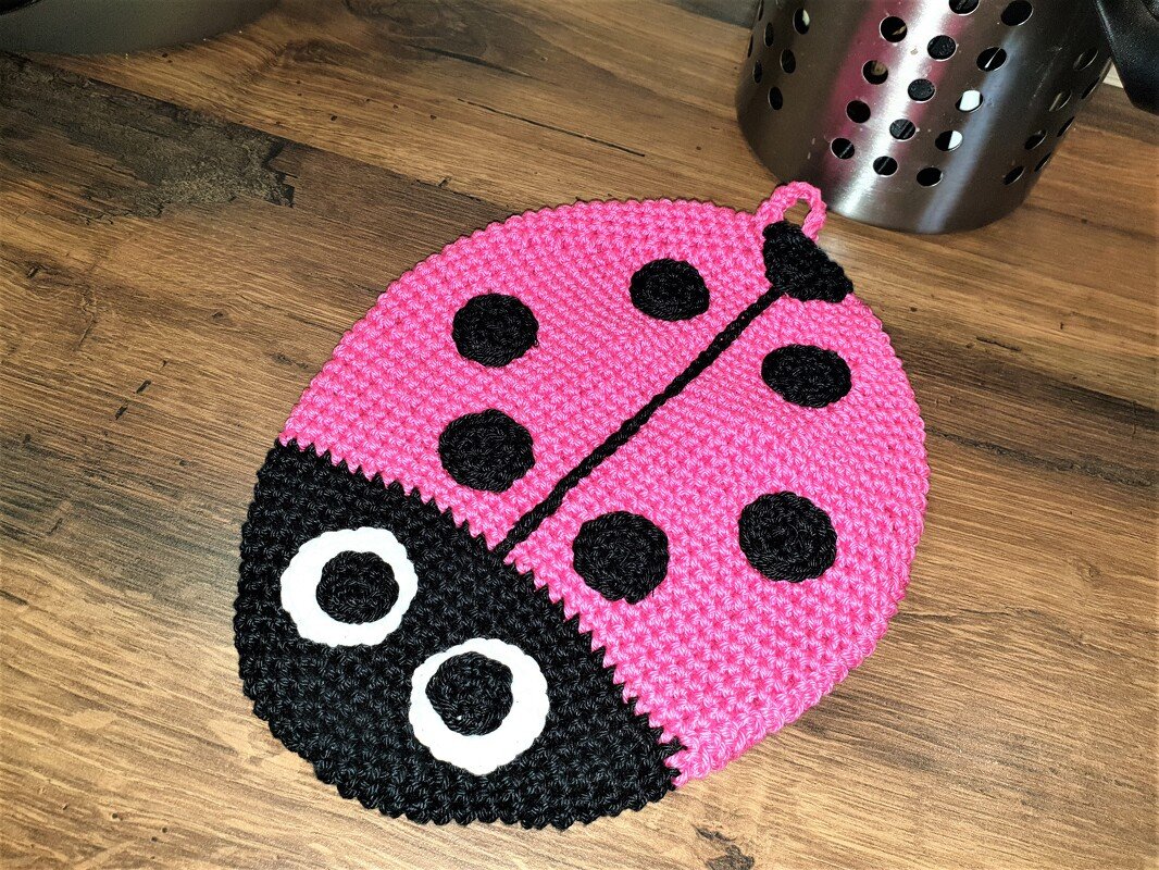 Gehäkelter pinker Marienkäfer-Topflappen mit schwarzen Punkten und weißen Augen auf Holz