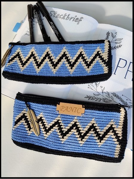 Zwei gehäkelte blaue Täschchen mit schwarzem und cremefarbenem Zickzackmuster, Federanhänger und Etikett mit Prägung PANIC