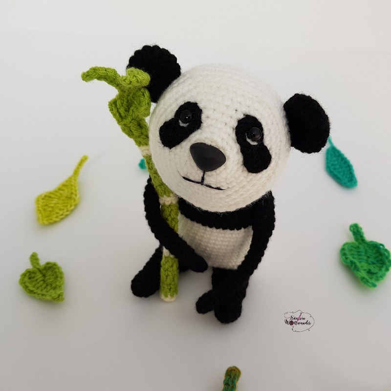 Gehäkelter Panda-Amigurumi in Weiß und Schwarz hält einen grünen gehäkelten Bambusstängel; umgeben von gehäkelten Blättern auf hellem Untergrund.