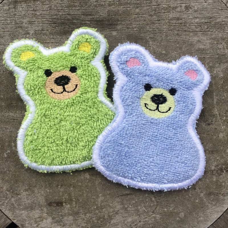 Zwei gestickte Bären-Applikationen in Frottee-Optik, grün und hellblau, mit weißen Konturen und bestickten Gesichtern auf Holzuntergrund