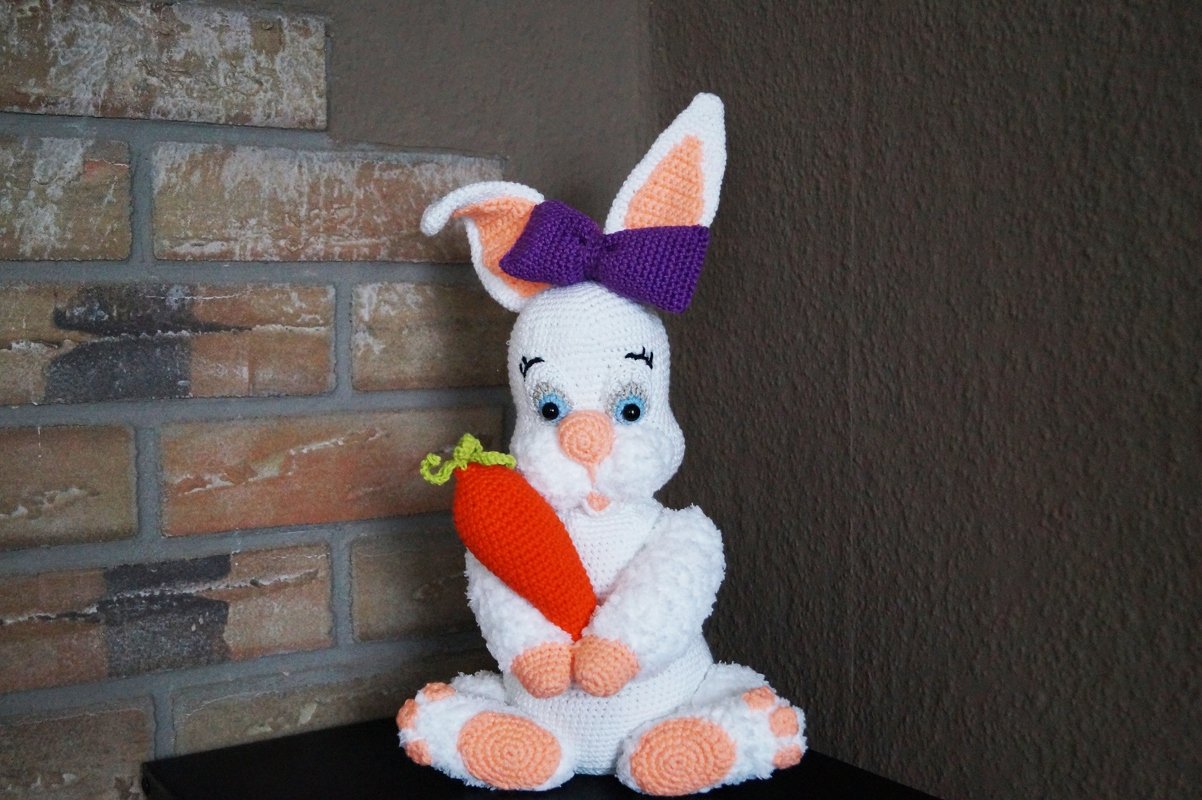Weiß gehäkelter Hase mit pfirsichfarbenen Pfoten, violetter Schleife und orangefarbener Möhre
