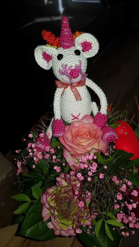 Weiße gehäkelte Einhorn-Maus mit pinken Details sitzt in einem Strauß rosa Rosen und Schleierkraut.