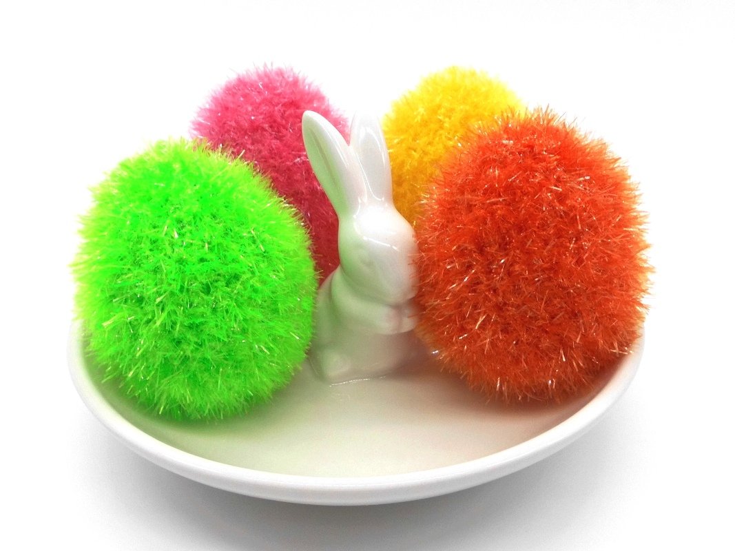 Weißer Teller mit weißer Keramik-Hasenfigur und vier flauschigen Häkel-Puscheln in Grün, Pink, Gelb und Orange.