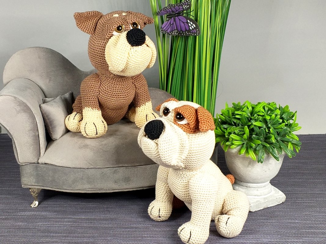 Two crocheted bulldog amigurumi toys: a brown-and-cream one on a small grey sofa and a cream-and-brown one on the floor beside plants.