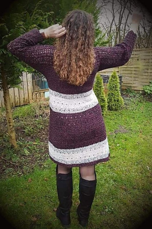 Rückenansicht: gehäkeltes dunkellila Kleid mit zwei weißen Querstreifen und hohen schwarzen Stiefeln im Garten