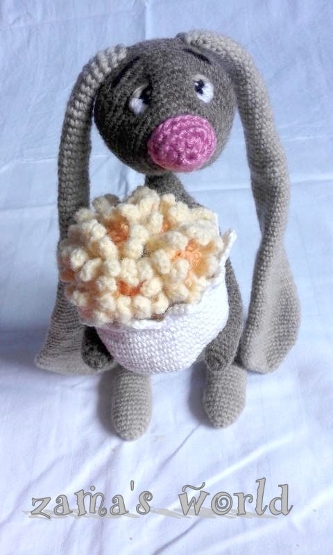 Gehäkelter grauer Amigurumi-Hase mit rosa Nase und langen Schlappohren, der einen weißen Korb mit creme- und apricotfarbenen Häkelblüten hält.