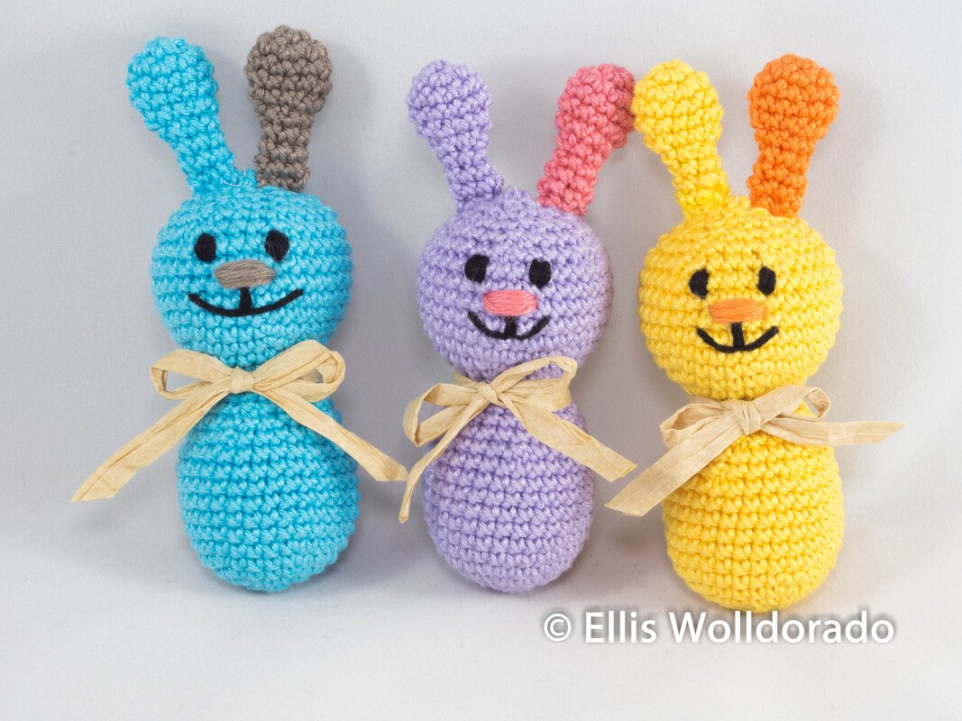 Drei bunte gehäkelte Hasenfiguren in Blau, Lila und Gelb mit Bastschleifen und aufgestickten Gesichtern.