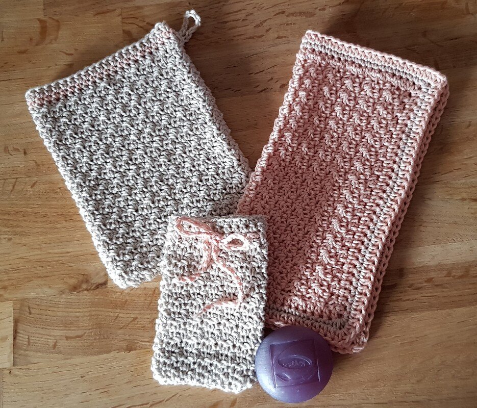 Zwei gehäkelte Tücher in Beige und Rosa, dazu ein gehäkelter Seifenbeutel mit Band und eine lila Seife auf einem Holztisch.