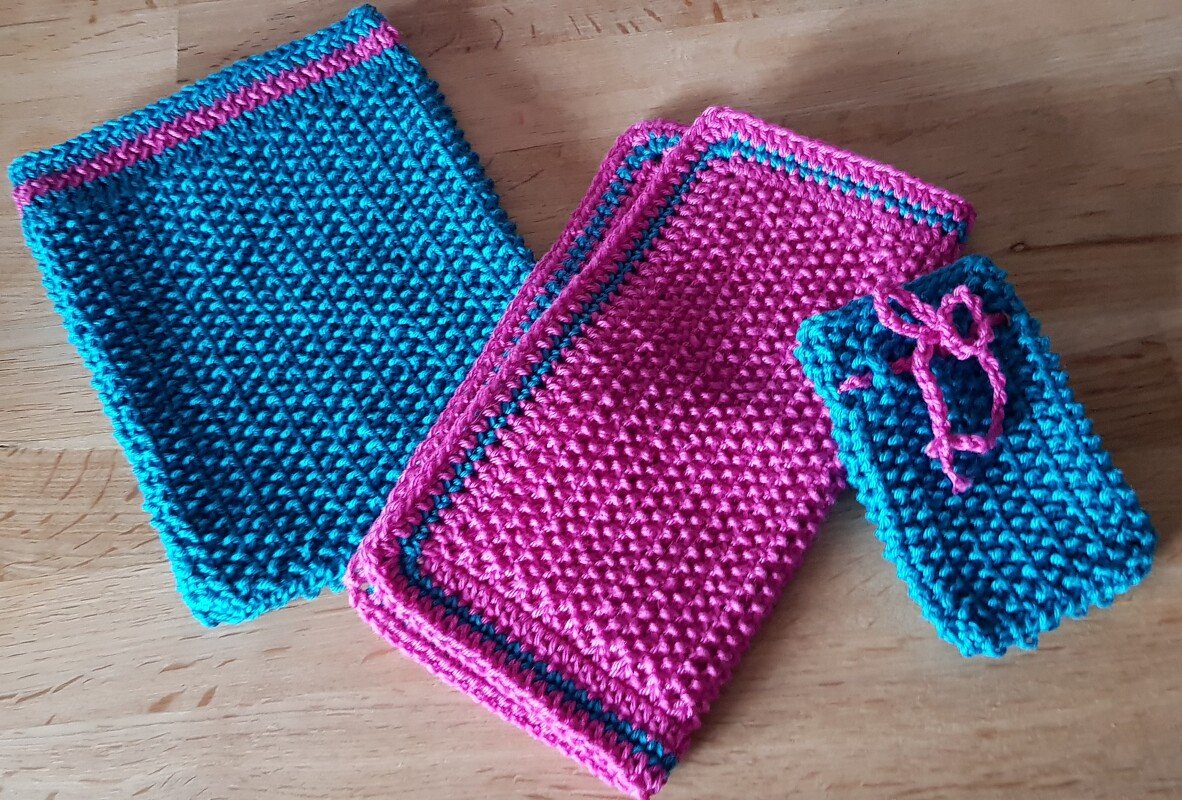 Zwei gestrickte Tücher in Pink, ein türkisfarbenes Tuch und ein kleiner türkisfarbener Beutel mit pinkem Band auf Holz.