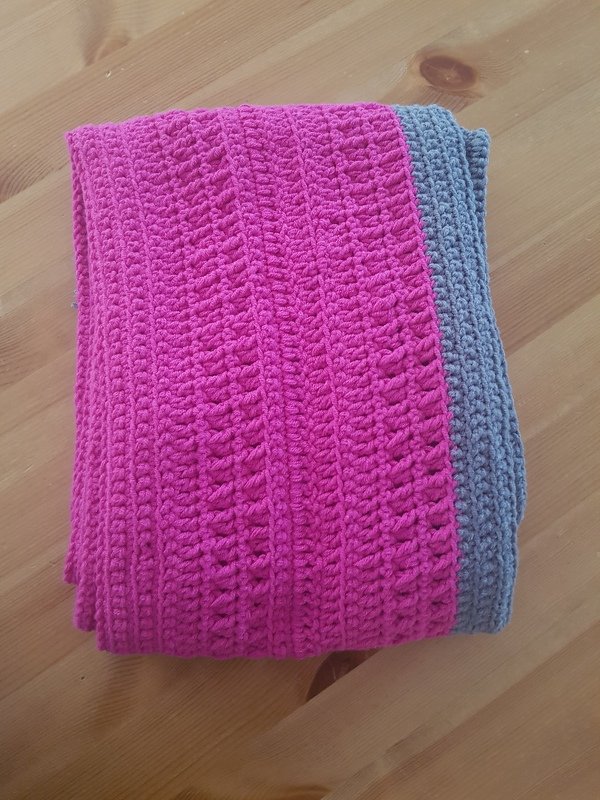 Gefalteter pinker Häkelschal mit grauer Kante und Reliefmuster auf Holztisch.