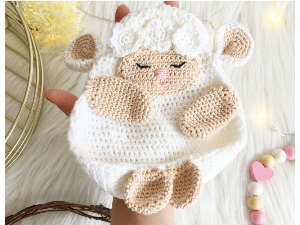 Gehäkeltes weiß-beiges Schaf-Schmusetuch mit schlafendem Gesicht, in einer Hand gehalten vor hellem Hintergrund.