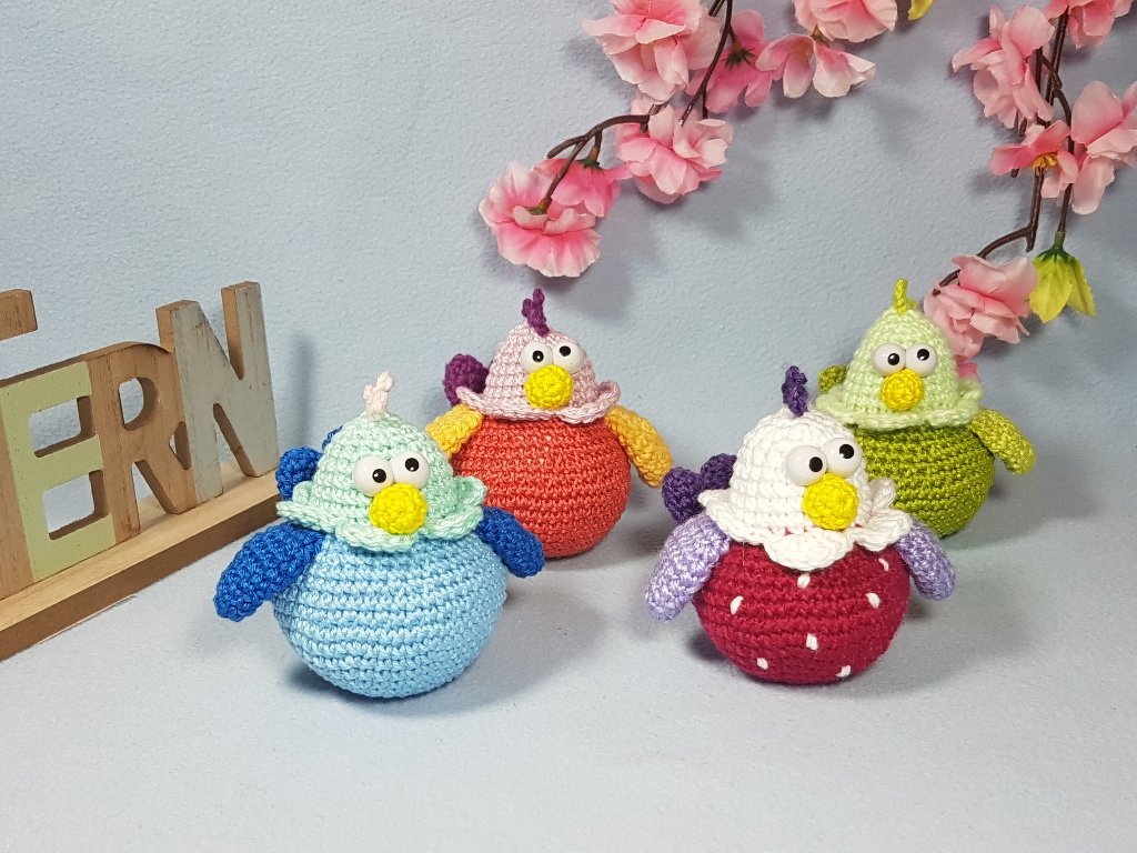 Vier gehäkelte Amigurumi-Küken in Blau, Orange, Rot und Grün mit großen Augen, umgeben von künstlichen Kirschblüten und Holzbuchstaben auf hellblauem Hintergrund.
