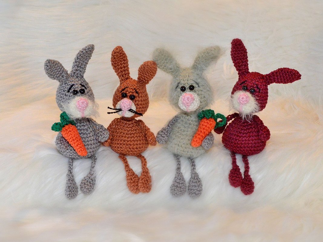 Vier gehäkelte Amigurumi-Hasen in verschiedenen Farben, zwei mit Karotten, auf weißem Flausch