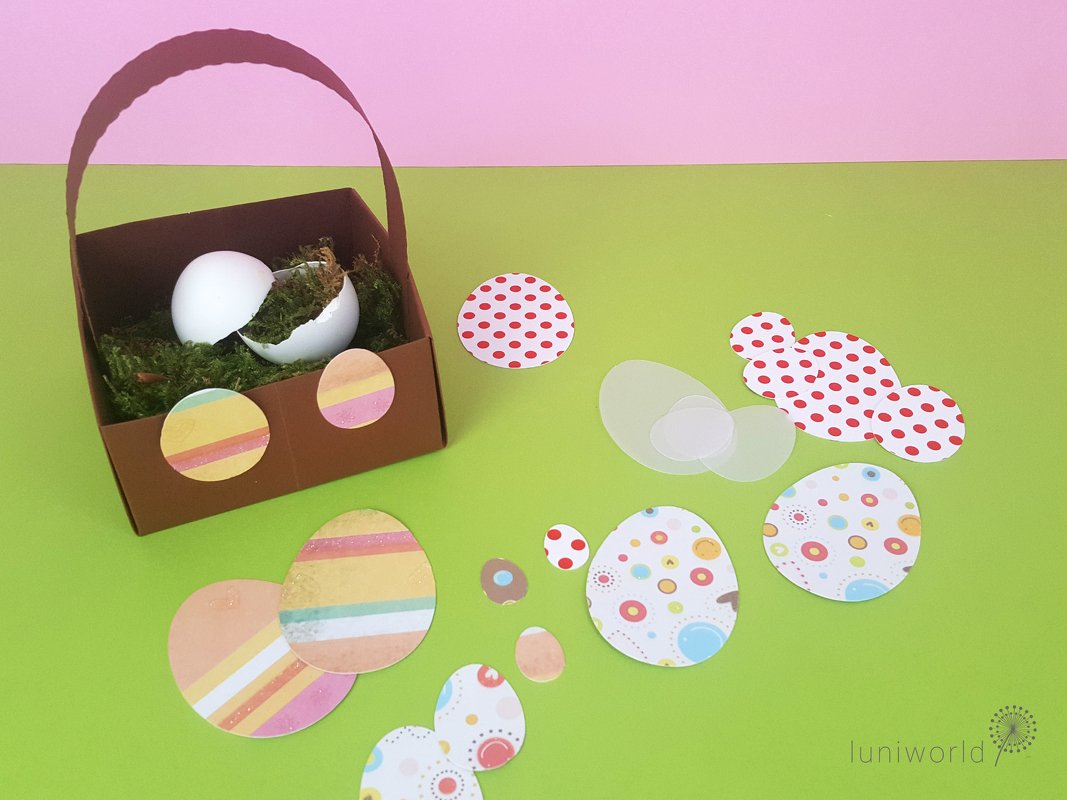 Braunes Papierkörbchen mit Moos und Eiern, umgeben von bunten ausgeschnittenen Papier-Ostereiern auf grünem Untergrund