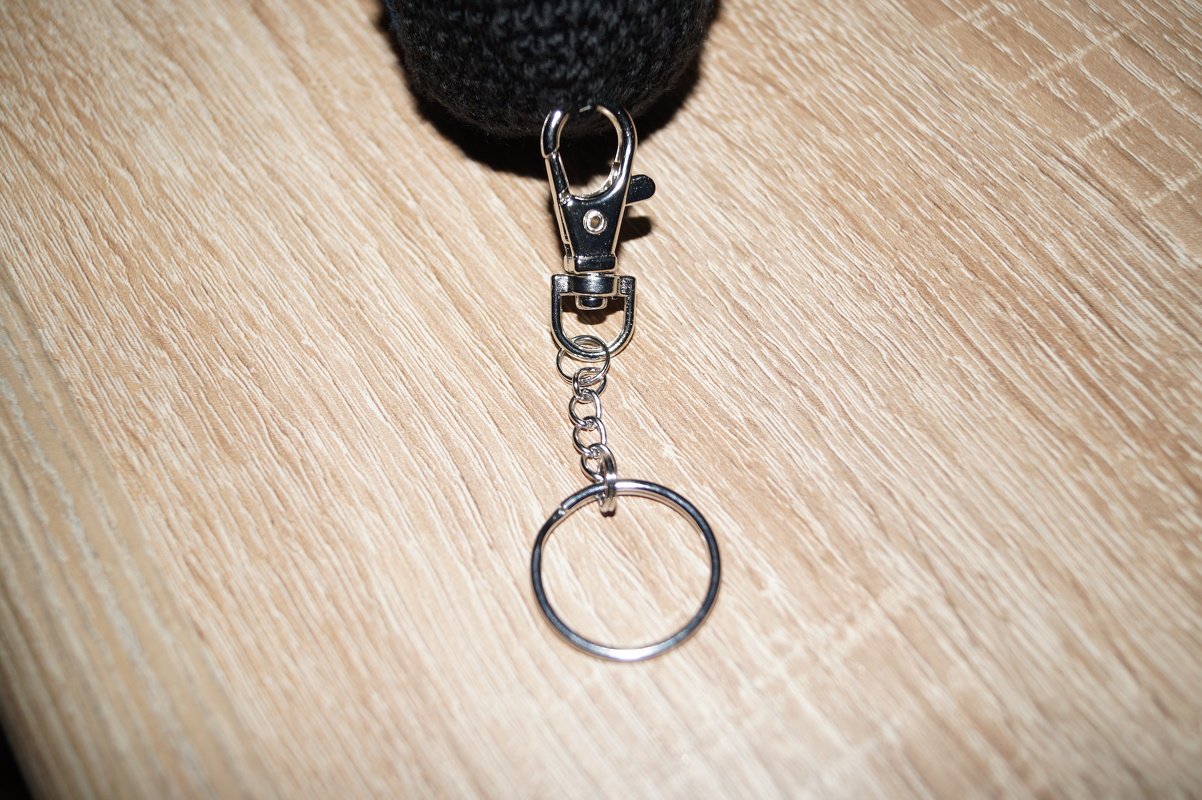 Dunkler gehäkelter Schlüsselanhänger mit silbernem Karabinerhaken und Schlüsselring auf hellem Holzuntergrund.