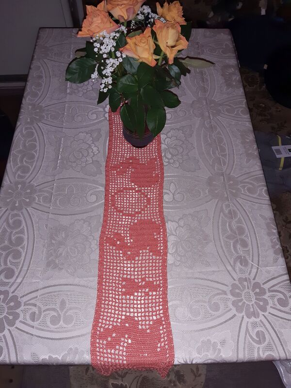 Lachsfarbener gehäkelter Tischläufer mit filigranem Muster, darauf ein Blumenstrauß aus orangen Rosen.