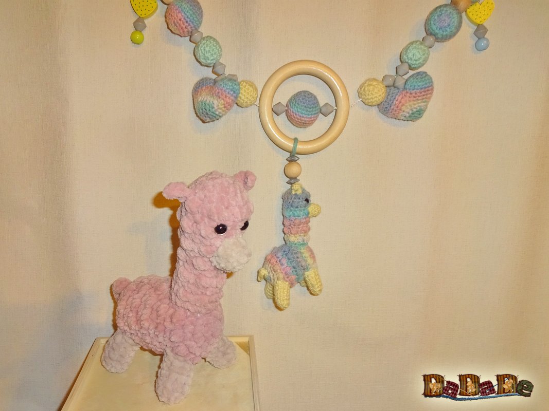 Rosa, flauschig gehäkeltes Lama-Plüschtier neben einem vielfarbigen Alpaka-Hängespielzeug an einem Holzring mit gehäkelten Perlen.